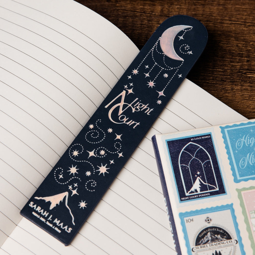A dark navy blue paper bookmark with celestial illustrations (moon, stars, constellation) and the words 'Night Court' and 'SARAH J MAAS' is placed on an open lined notebook. A matching navy blue zippered pouch is partially visible at the top, and a sheet of decorative stickers is on the right, all on a wooden surface.