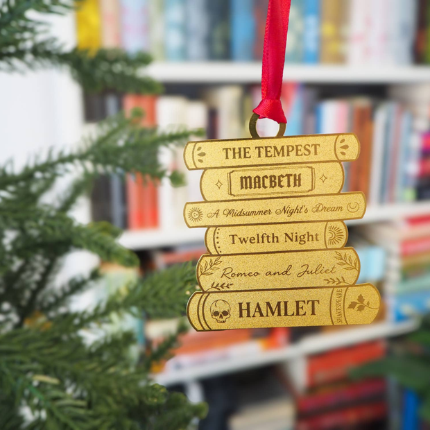 A stack of Shakespeare plays etched into a brass book stack ornament with a red ribbon hanging from a blurred out Christmas tree with bookshelves in the background.