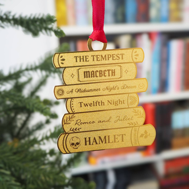 A stack of Shakespeare plays etched into a brass book stack ornament with a red ribbon hanging from a blurred out Christmas tree with bookshelves in the background.