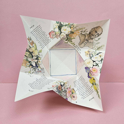 A square origami paper keepsake with illustrated floral and portrait designs, photographed on a solid pink background.