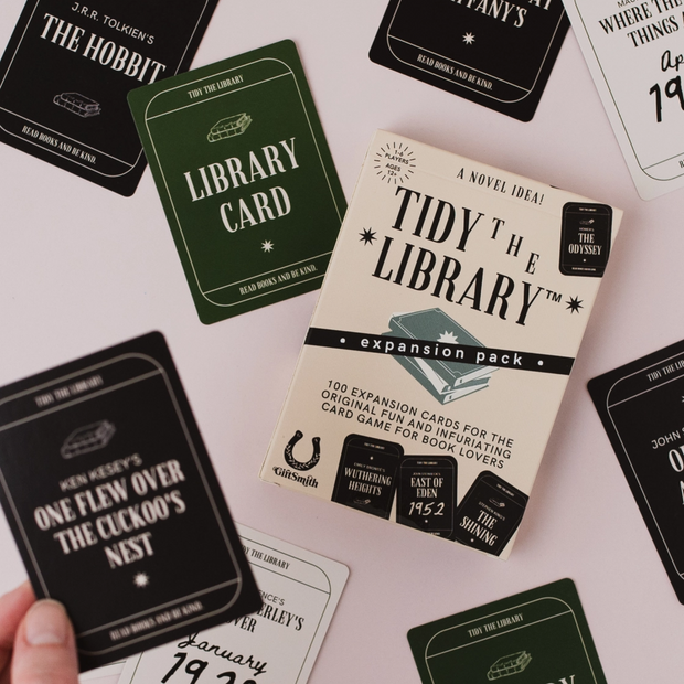 A boxed card game called "Tidy the Library" expansion pack sits on a pale pink surface, surrounded by several sample cards in black, green, and white with book-themed quotes and titles. A hand is holding one black card at the lower left.