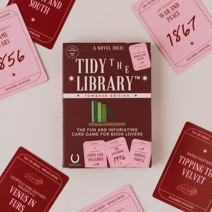 The 'Tidy the Library: Romance Edition' card game box is centered on a light beige background, surrounded by scattered pink and dark red cards showing different historical romance titles and years.