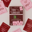 The 'Tidy the Library: Romance Edition' card game box is centered on a light beige background, surrounded by scattered pink and dark red cards showing different historical romance titles and years.