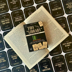 The image shows the card game 'Tidy the Library' laid out on an open book. The game includes a rulebook, library cards, and book cards with various book titles.