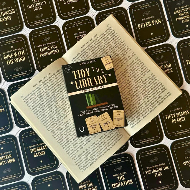 The image shows the card game 'Tidy the Library' laid out on an open book. The game includes a rulebook, library cards, and book cards with various book titles.