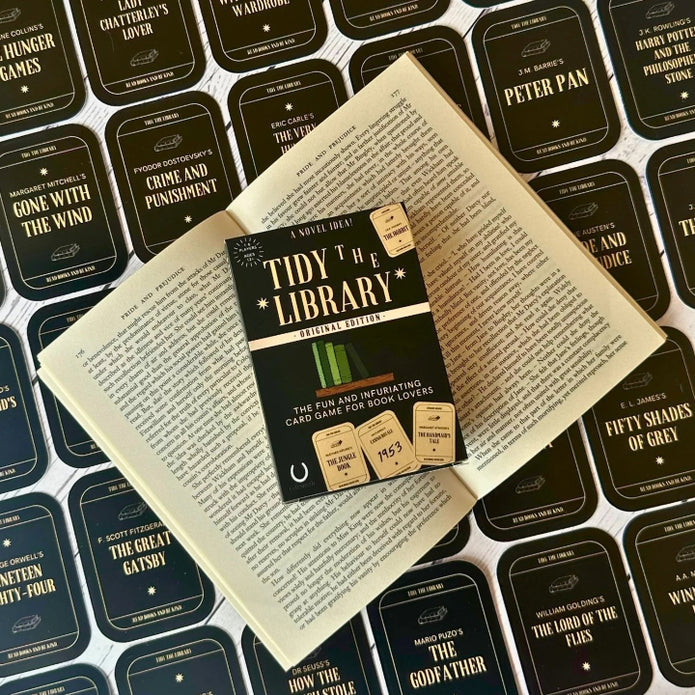 The image shows the card game 'Tidy the Library' laid out on an open book. The game includes a rulebook, library cards, and book cards with various book titles.