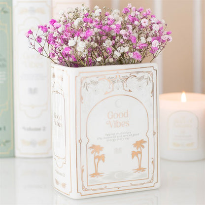 Decorative ceramic book shaped vase with 'Good Vibes' text and pink flowers on a light background