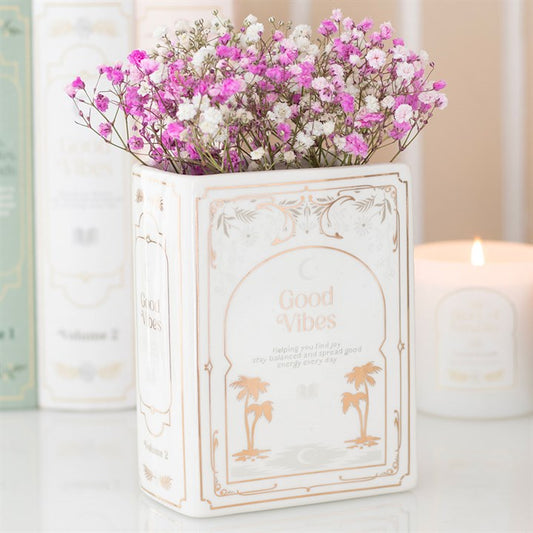Decorative ceramic book shaped vase with 'Good Vibes' text and pink flowers on a light background