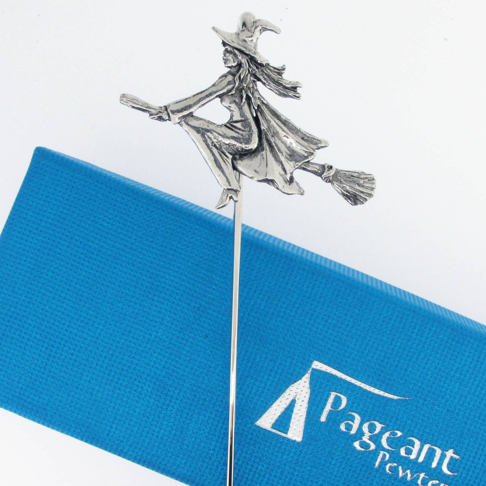 A silver-toned metal witch riding a broomstick pin or decoration, displayed above a blue branded box with the text 'Pageant Powter'.