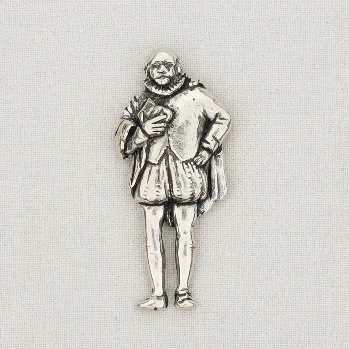 A pewter metal pin depicting a William Shakespeare in a ruff collar and knee-length tunic, shown on al ight textured fabric background.